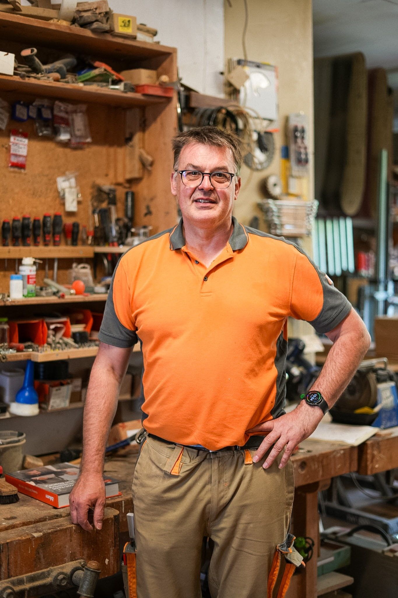 Jörg Häfner steht in Arbeitskleidung an seiner Hobelbank, eine Hand aufgestützt, hinter ihm ein Regal voller Werkzeug.